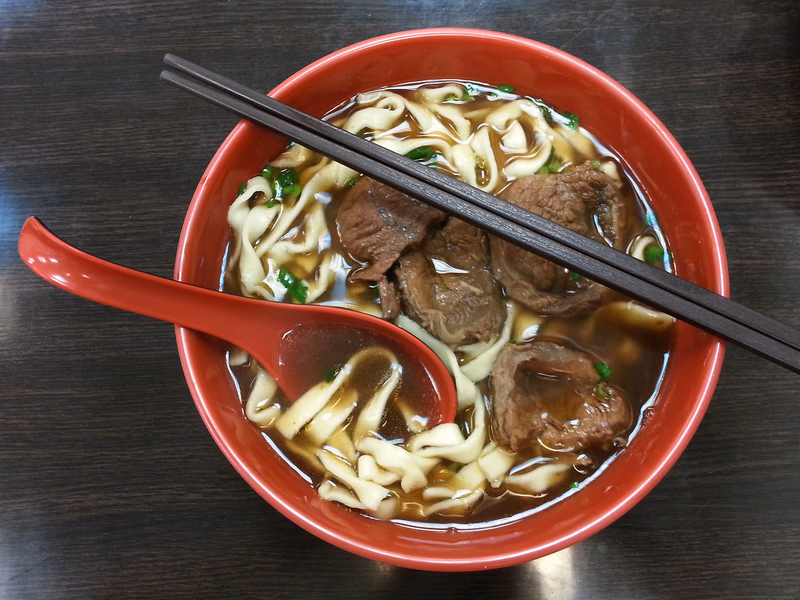 A bowl of beef noodle soup with chopsticks and a red spoon